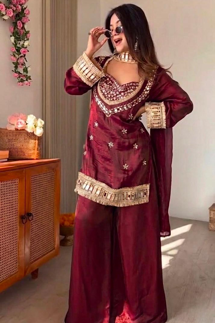 Woman in a maroon traditional outfit with gold accents in a room with a wooden cabinet and floral decorations.