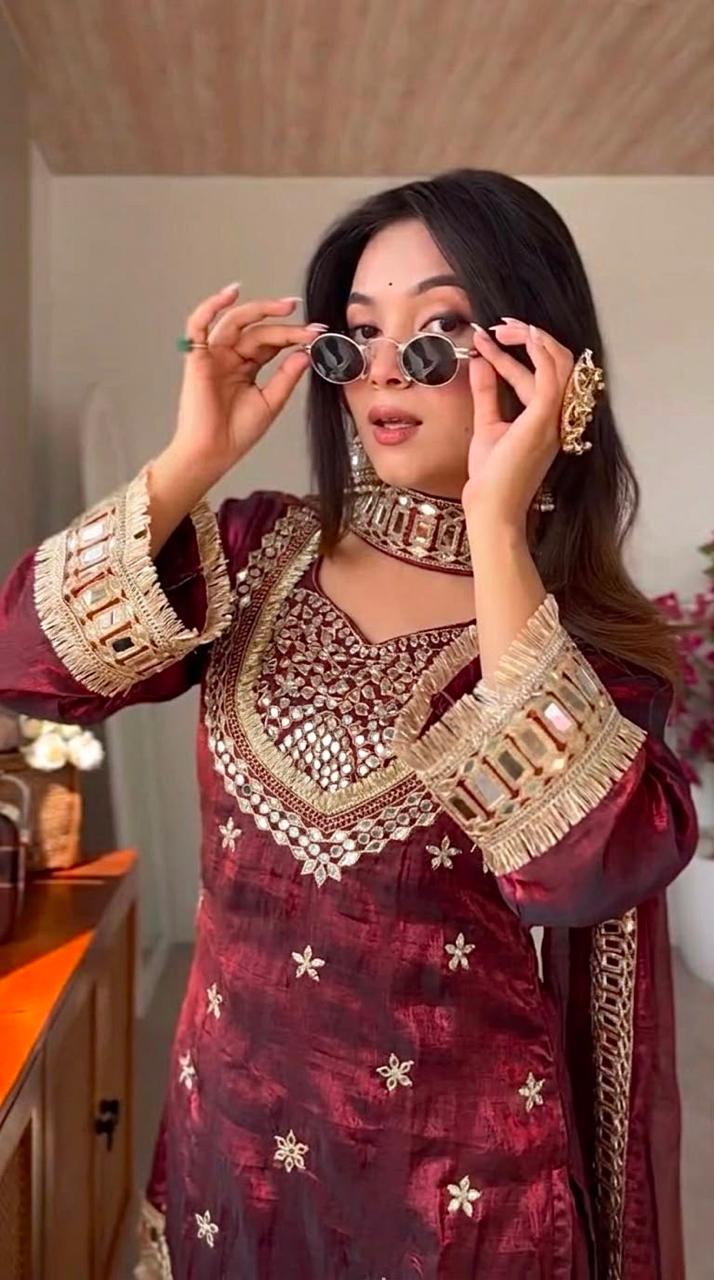 Woman in traditional maroon and gold outfit adjusting eyewear in a room.