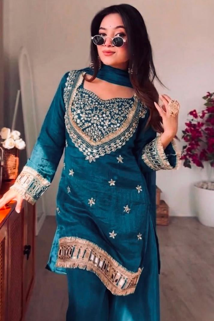 Woman wearing a teal traditional outfit with intricate patterns in a room.