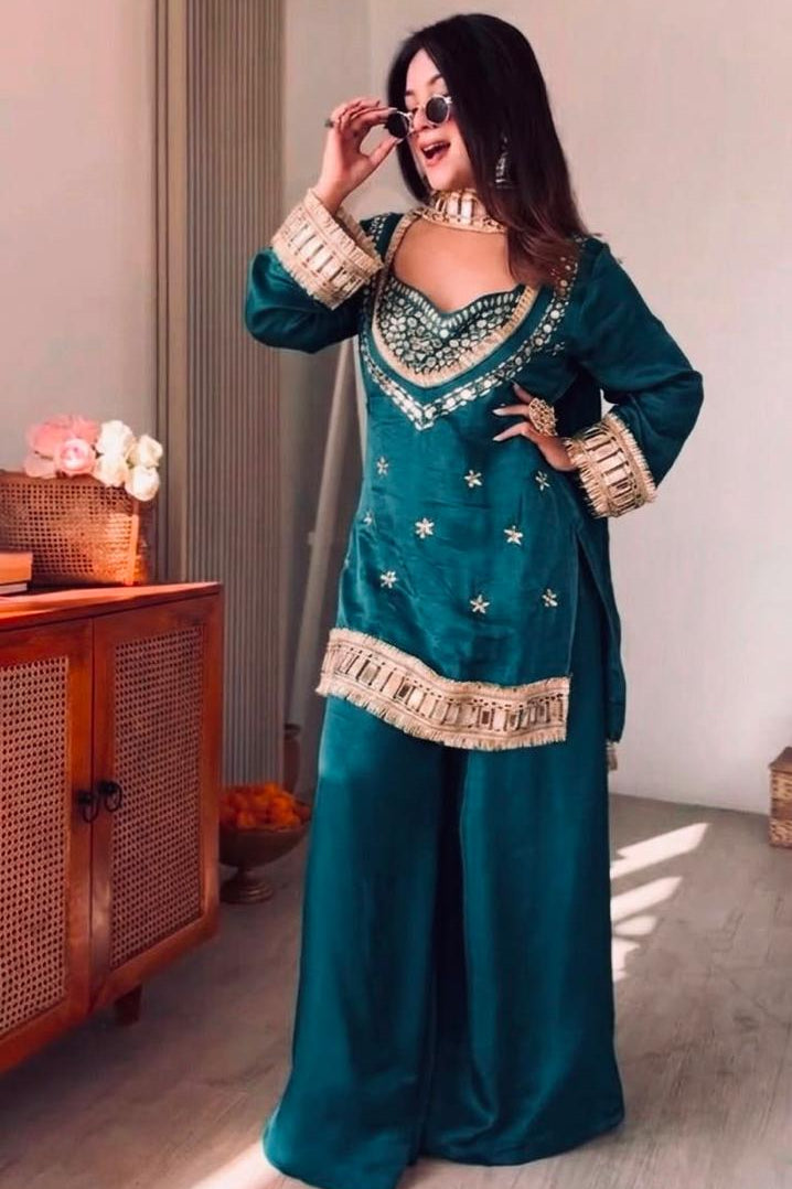 Woman in a teal traditional outfit with gold accents standing in a room with wooden furniture.
