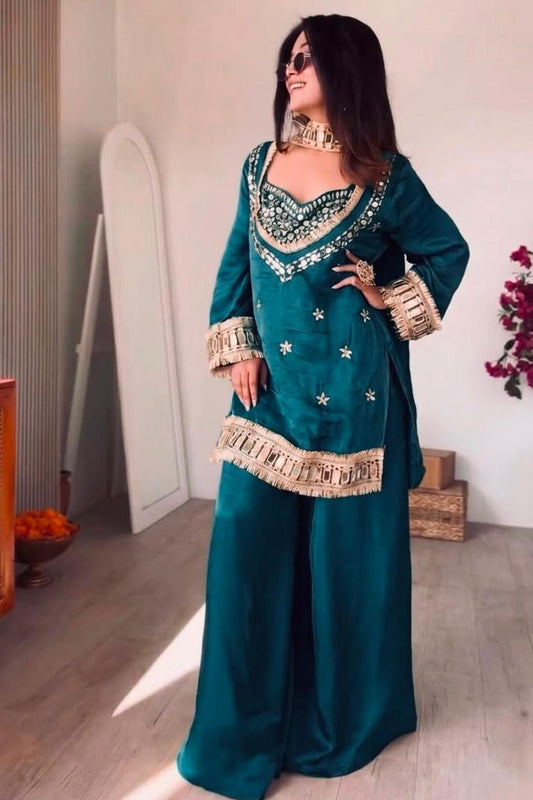 Woman in a teal traditional outfit with gold embellishments standing indoors.
